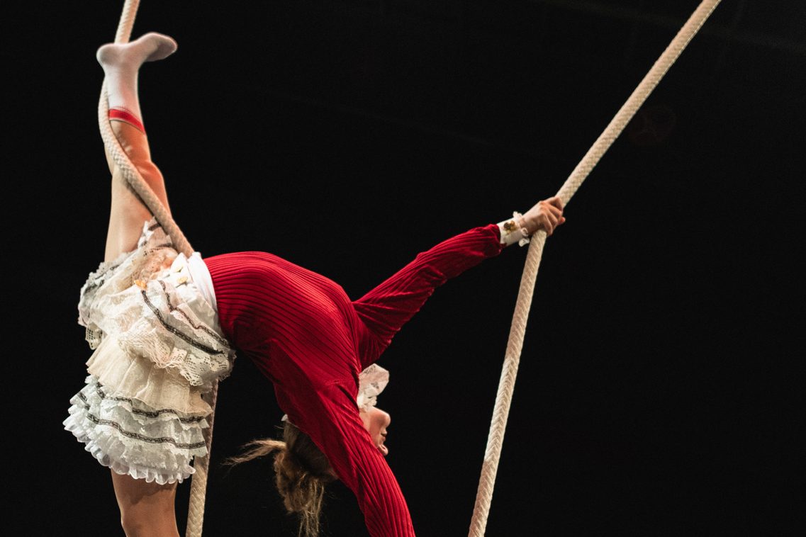 Eine Frau in vertikaler Spagat-Position zeigt am Seil eine Rückbeuge in der Luft – Kunst des Schwungseils & Clownerie