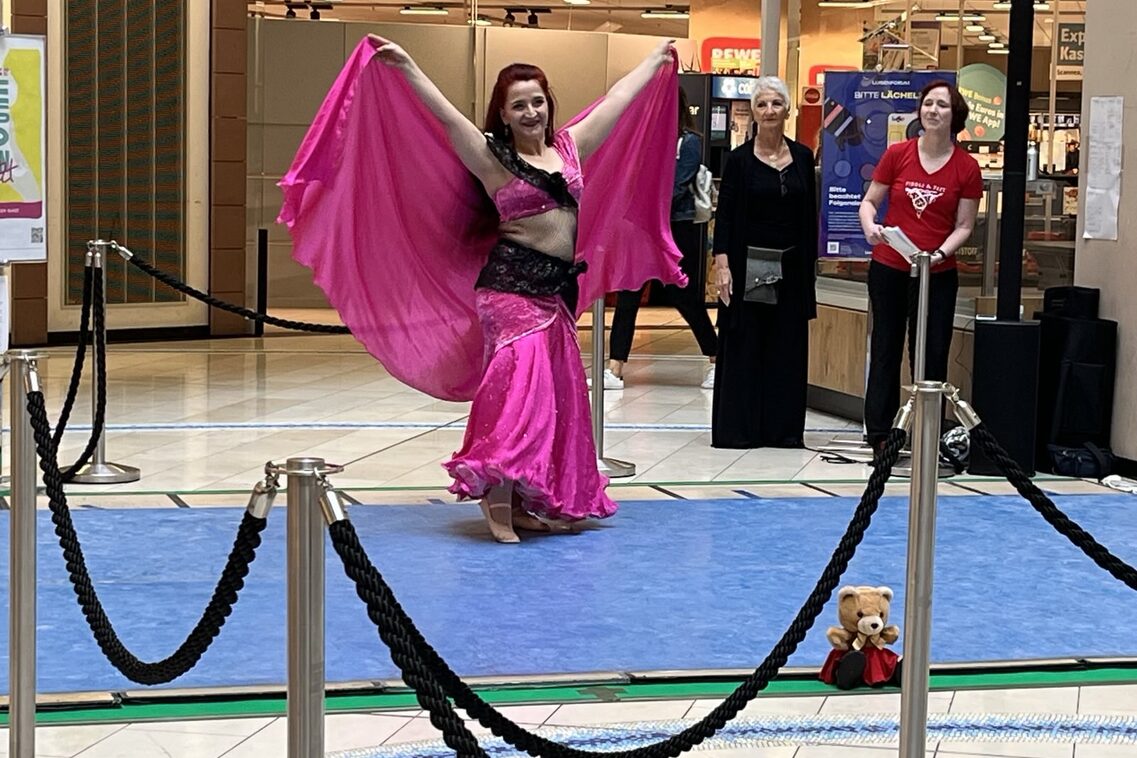 Eine Bauchtänzerin in lila-schwarzem Kostüm hebt beide Arme und breitet ein großes Tuch aus.