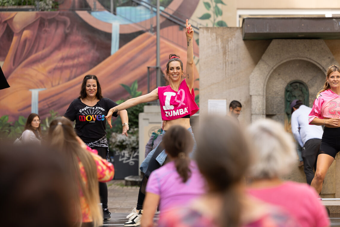 Eine Zumbatrainerin steht vor einer Gruppe, hält einen Arm hoch und lächelt.