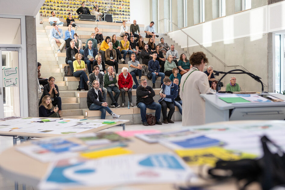 Eine Veranstaltung in einem modernen, offenen Gebäude mit hohen Fenstern und einer breiten Sitztreppe. Eine große Gruppe von Menschen sitzt auf den Stufen, während eine Person im Vordergrund an einem Rednerpult spricht. Auf den Tischen vor der Rednerin liegen verschiedene Broschüren, Zettel und Arbeitsmaterialien. Die Atmosphäre wirkt konzentriert und partizipativ.