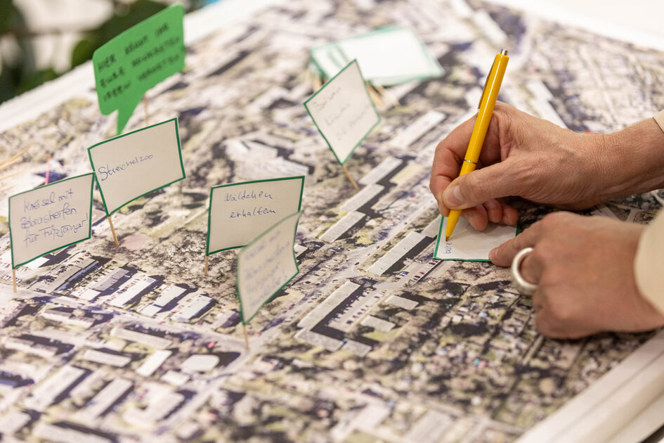 Eine Nahaufnahme einer Hand mit einem silbernen Ring, die mit einem gelben Stift auf eine kleine Karte schreibt. Die Karte wird auf eine große Stadtkarte gelegt, auf der bereits mehrere andere kleine Kärtchen mit handschriftlichen Notizen und grünen Sprechblasen angebracht sind. Die Notizen enthalten Ideen oder Vorschläge für die Stadtentwicklung, zum Beispiel „Wäldchen erhalten“ und „Streichelzoo“.
