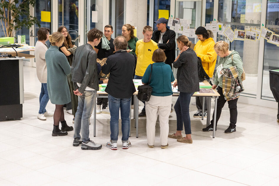 Eine Gruppe von Menschen steht um einen Tisch in einem lichtdurchfluteten Raum. Sie diskutieren und schauen sich gemeinsam Karten und Dokumente an. Einige tragen gelbe Hoodies, die möglicherweise eine Zugehörigkeit zu einer Organisation oder einem Team anzeigen. Im Hintergrund hängen an einer Glaswand Informationsmaterialien und Fotos. Die Szene wirkt interaktiv und gemeinschaftlich.