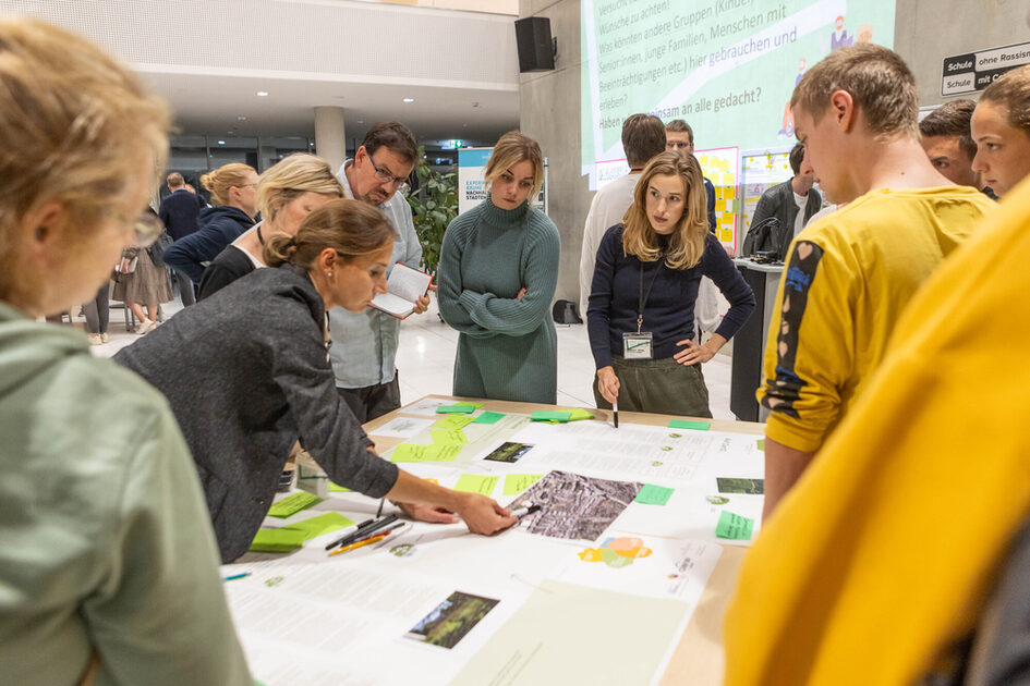 Eine Gruppe von Menschen steht um einen Tisch und betrachtet große Plakate mit Karten, Texten und grünen Notizzetteln. Eine Frau im dunklen Blazer zeigt auf eine Stelle auf dem Papier, während andere aufmerksam zuhören. Eine Frau mit Namensschild hält einen Stift und scheint etwas zu erklären. Im Hintergrund sind weitere Personen in einer hell beleuchteten Veranstaltungshalle.