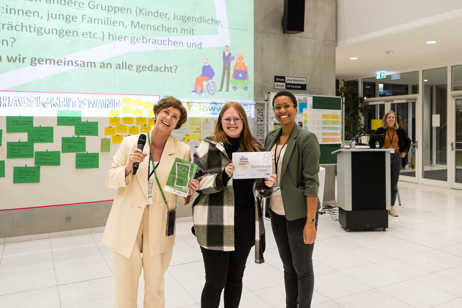 Drei Frauen stehen lächelnd in einer Veranstaltungshalle. Die Frau links trägt einen hellen Anzug und hält ein Mikrofon sowie einen grünen Flyer. Die Frau in der Mitte trägt eine karierte Jacke und hält ein Deutschlandticket. Die Frau rechts trägt einen grünen Blazer und hat ebenfalls ein Namensschild. Im Hintergrund ist die Nachhaltigkeitswand mit grünen und gelben Kärtchen zu sehen.