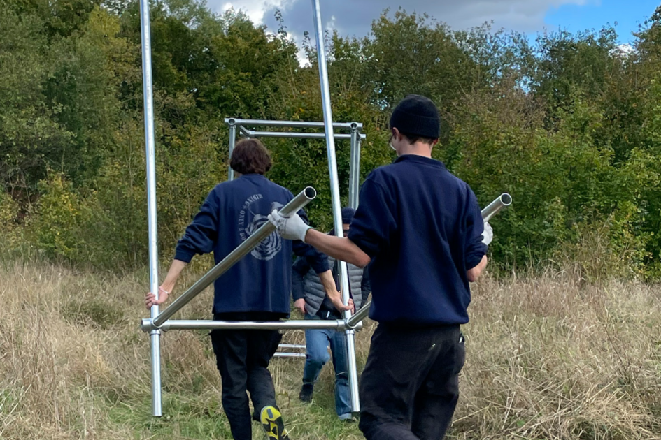 Zwei junge Männer in dunkler Kleidung tragen metallene Gerüstelemente auf eine verwilderte Wiese.