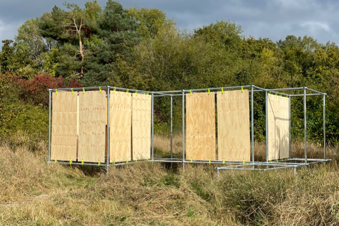 Eine einfache, rechteckige Struktur aus Metallrohren steht inmitten einer Wiese. Die Wände bestehen aus hellen Sperrholzplatten, die mit gelben Kabelbindern an der Metallkonstruktion befestigt sind. Die Struktur ist teilweise offen, sodass man hindurchsehen kann. Die Umgebung ist eine natürliche Landschaft mit hohem, trockenem Gras und vereinzelten Bäumen im Hintergrund. Der Himmel ist bewölkt mit dunklen Wolken, durch die stellenweise Licht fällt.