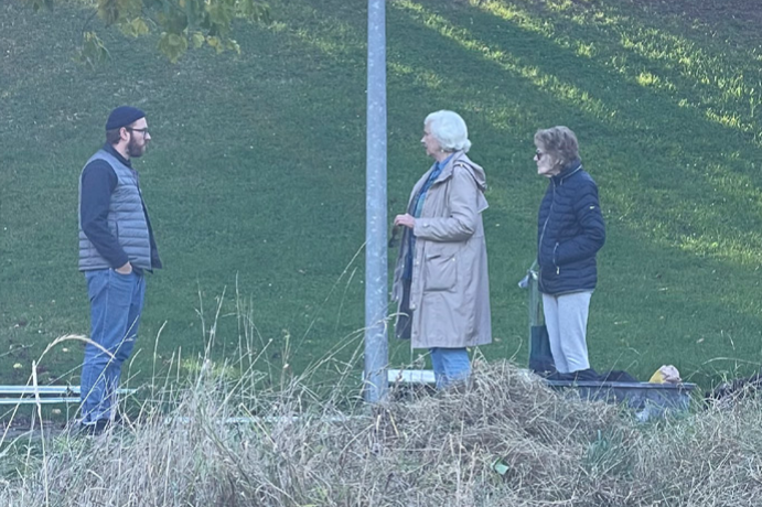 Ein Mann mit herbstlicher, dunkler Kleidung und zwei ältere Frauen stehen sich gegenüber und unterhalten sich. Sie werden von weitem beobachtet, im Vordergrund vertrocknetes Heidegras. Eine Laterne teilt das Bild mittig.