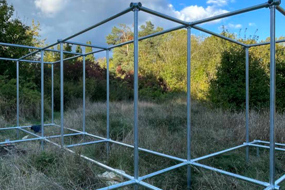 Eine rechteckige, offene Metallstruktur aus Rohren steht in einer wilden, grasbewachsenen Landschaft. Die Konstruktion bildet ein Gerüst mit mehreren gleichmäßig verteilten, würfelförmigen Segmenten. Der Himmel ist blau mit weißen Wolken, und im Hintergrund sind Bäume und Sträucher zu sehen.