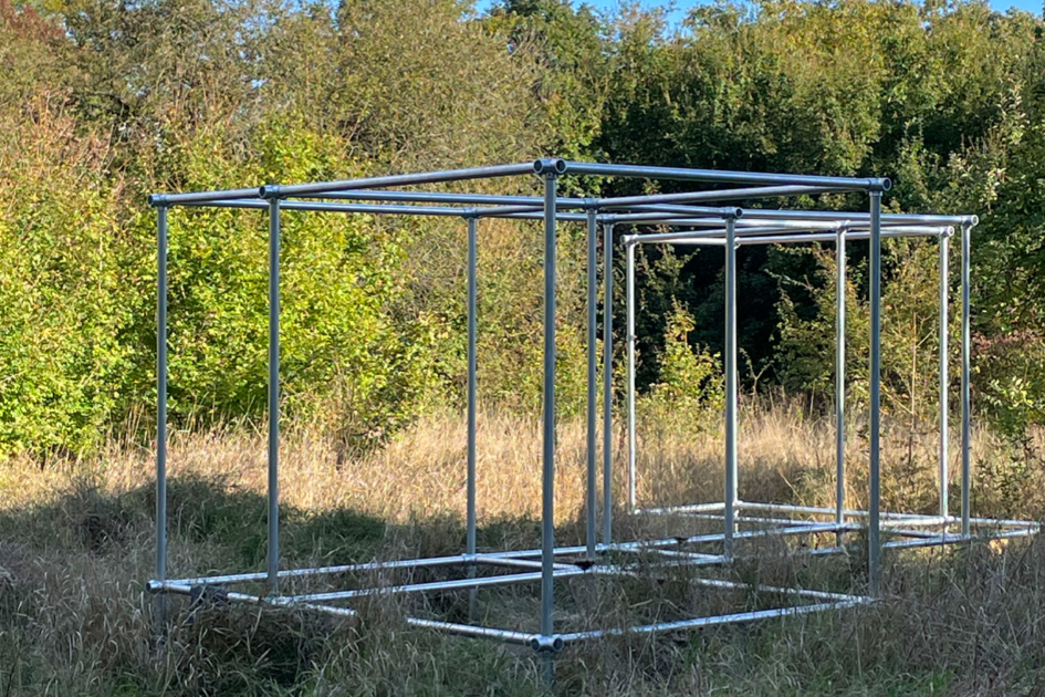 Eine rechteckige, offene Metallstruktur aus Rohren steht in einer wilden, grasbewachsenen Landschaft. Die Konstruktion bildet ein Gerüst mit mehreren gleichmäßig verteilten, würfelförmigen Segmenten. Der Himmel ist blau mit weißen Wolken, und im Hintergrund sind Bäume und Sträucher zu sehen.