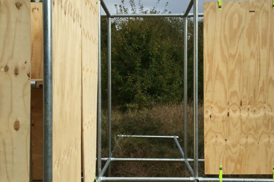Eine Metallgerüst-Konstruktion mit befestigten Holzplatten an den Seiten. Die Platten sind mit gelben Halterungen an den Metallrohren fixiert. Ein gepflasterter Bereich führt in die Struktur hinein, während im Hintergrund hohes Gras und Sträucher wachsen. Der Himmel ist teils bewölkt.