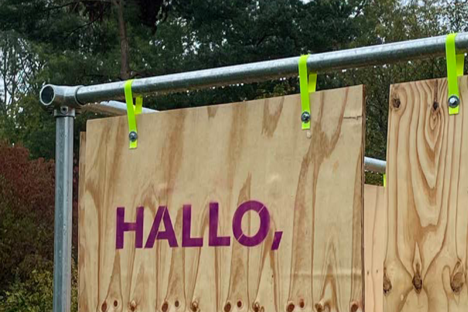 Das Bild zeigt eine Holzplatte mit der Aufschrift „HALLO,“ in lila Buchstaben. Die Holzplatte ist mit leuchtend gelben Befestigungen an einem Metallgerüst angebracht. Im Hintergrund sind Bäume und ein bewölkter Himmel zu sehen. Das Holz hat eine natürliche Maserung mit Astlöchern.