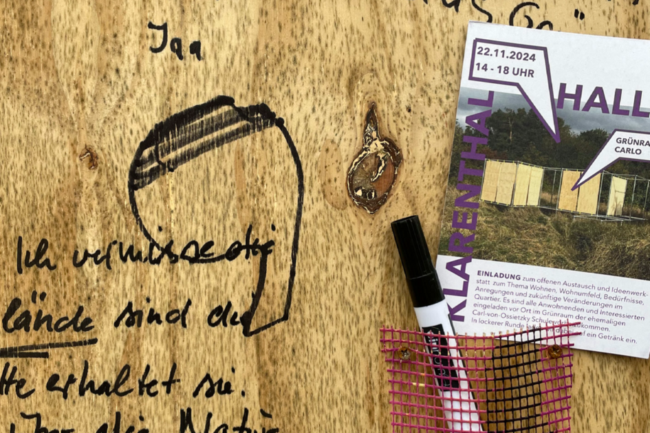 Nahaufnahme einer Sperrholzplatte, auf die mit schwarzem Filzstift in verschiedenen Handschriften Kommentare geschrieben wurden. Lesbar ist „bitte keine Wohnungen bauen wir lieben das Grün“. An der Sperrholzplatte ist ein schwarzer Filzstift mit Hilfe eines Plastiknetztes befestigt. Daneben hängt ein Flyer, auf dem die Sperrholzplattenkonstruktion abgebildet ist und auf dem als Überschrift das Wort „Hallo“ prankt.