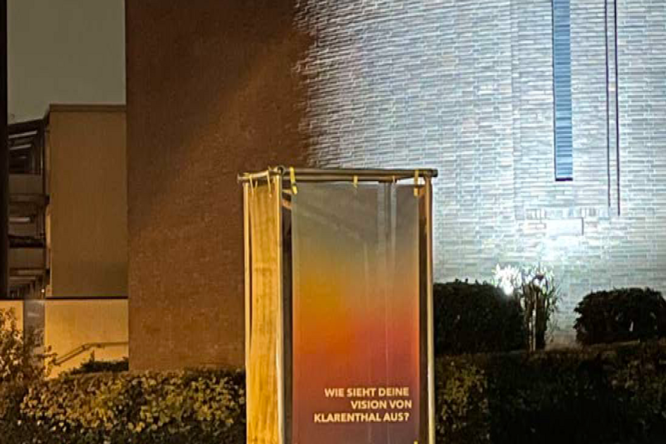 Eine Metallrahmenkonstruktion mit einem daran befestigten Banner steht an einer Straßenecke. Es ist im dunkel und im Hintergrund ist eine beleuchtete Fassade eines aus dunklen Backsteinen gebauten Gebäudes zu sehen.