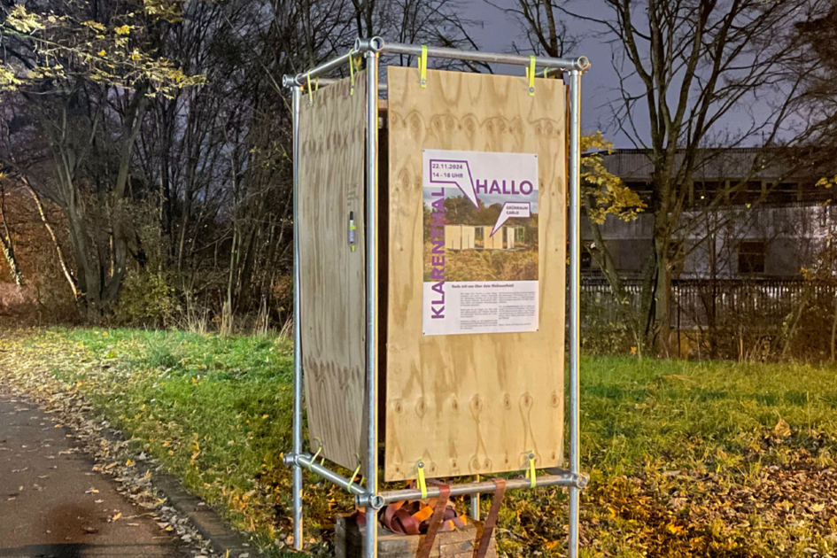 Eine Metallrahmenkonstruktion mit einer daran befestigten Holzplatte steht an einer Weggabelung auf grünem Rasen. Es liegen viele orangene Blätter auf dem Boden. Es ist im dunkel und im Hintergrund sind Bäume. Die Konstruktion wird angestrahlt und auf der Holzplatte ist ein Plakat befestigt.
