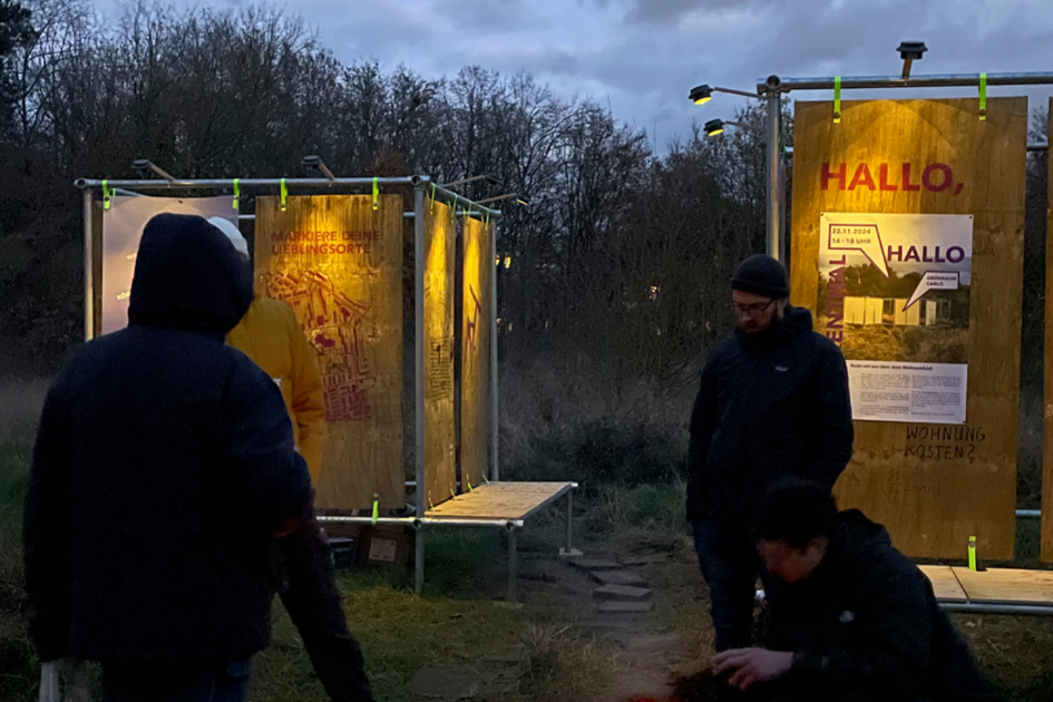 Es dämmert und im Vordergrund des Bildes sind die Silhouetten dreier Personen zu erkennen, von denen sich eine neben einer Feuerschalte hingehockt hat. Im Hintergrund ist eine Metallkonstruktion mit daran befestigten Sperrholzplatten zu erkennen. Diese sind jeweils beleuchtet und an ihnen sind teilweise Plakate befestigt, teilweise sind die Platten auch beschriftet.
