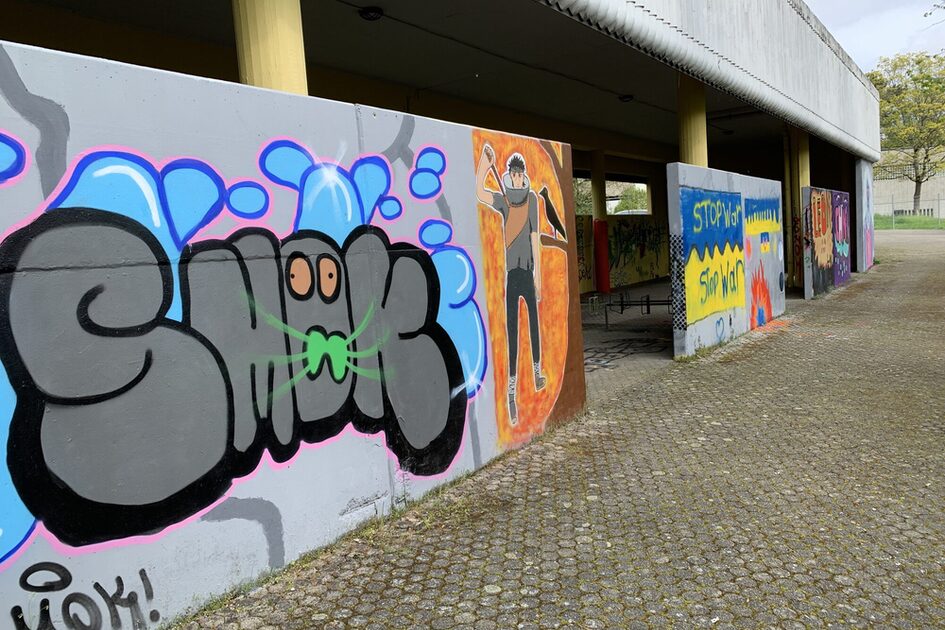 Das Bild zeigt eine mit Graffiti verzierte Betonwand in einem überdachten Außenbereich. Der Boden ist mit runden Pflastersteinen belegt, und im Hintergrund sind eine Wiese, Bäume und ein Zaun zu sehen. Graffiti-Details: •	Links ist ein großes Graffiti-Wort in grauen, dicken Buchstaben mit schwarzer Umrandung zu sehen. Die Buchstaben haben zwei orangefarbene Augen und grüne Linien, die an Schnurrhaare erinnern. Blaue Blasen umgeben das Wort, und ein pinker Rand hebt es von der Wand ab. •	In der Mitte ist eine Figur auf einer orangefarbenen Fläche gesprüht. Sie trägt dunkle Kleidung, hat ein Stirnband und hebt beide Hände vors Gesicht. •	Weiter rechts sind verschiedene kleinere Graffiti zu erkennen, darunter der Schriftzug "STOP WAR" in Blau und Gelb, sowie weitere bunte Designs mit Feuer- und Muster-Elementen. •	Die Wand gehört zu einem Gebäude mit gelben Säulen und einem grauen Wellblechdach, das Schutz vor Regen bietet.