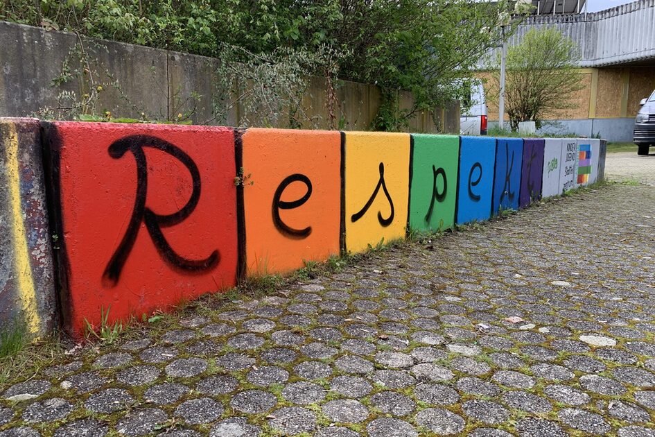 Das Bild zeigt eine Reihe von rechteckigen Betonblöcken, die als Begrenzung oder Absperrung dienen. Jeder Block ist in einer anderen Farbe des Regenbogens bemalt: Rot, Orange, Gelb, Grün, Blau und Violett. Zusammen bilden die Blöcke das Wort „Respekt“, das in schwarzer, handgeschriebener Schrift darauf gesprüht wurde. Rechts neben dem letzten Buchstaben befinden sich kleinere Schriftzüge und ein Regenbogenmotiv. Die Blöcke stehen auf einem gepflasterten Weg mit rundlichen Steinen, zwischen denen etwas Moos wächst. Im Hintergrund sind eine Betonmauer mit Pflanzen, ein Parkplatz mit einem Auto sowie ein Gebäude mit Wellblechdach und teils verbarrikadierten Fenstern zu sehen. Der Himmel ist bewölkt.