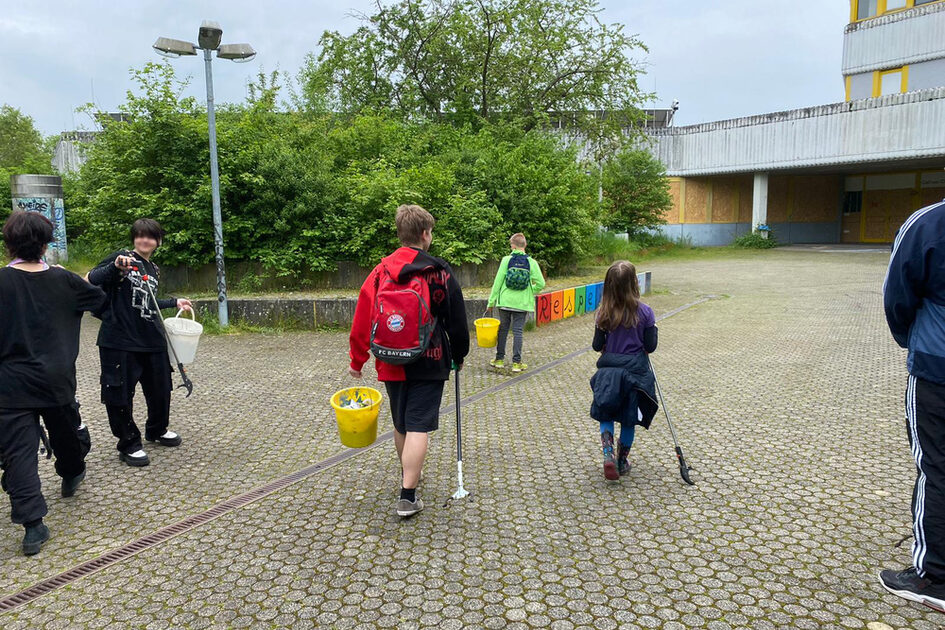 Eine Gruppe von Kindern und Jugendlichen läuft über einen gepflasterten Platz. Sie tragen Müllzangen und Eimer, in denen sie gesammelten Müll aufbewahren. Im Hintergrund ist ein Gebäude mit gelben und grauen Elementen zu sehen, teilweise mit Holzplatten verbarrikadiert. Eine bunte Sitzbank mit der Aufschrift „RESPEKT“ steht vor einer grünen Hecke.
