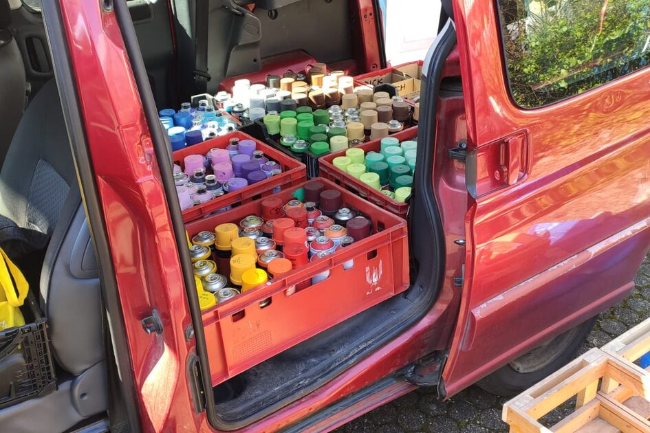 Das Bild zeigt die offene Schiebetür eines roten Transporters oder Vans, der auf einem gepflasterten Boden mit rundlichen Steinen steht. Im Inneren des Fahrzeugs befinden sich mehrere rote Kunststoffkisten, die randvoll mit Sprühdosen in verschiedenen Farben gefüllt sind. Die Farbdosen sind ordentlich sortiert, mit Deckeln in Gelb, Rot, Grün, Blau, Braun und Grau. Hinter dem Fahrzeug ist eine blaue mobile Toilette mit der Aufschrift "TOI TOI" sichtbar, sowie eine mit Brettern verbarrikadierte Wand oder Tür. Rechts im Vordergrund befinden sich gestapelte Holzkisten oder Paletten. Das Bild wurde bei Sonnenschein aufgenommen, da Sonnenstrahlen und Schatten sichtbar sind. Die offene Tür des Fahrzeugs gibt den Blick auf den Innenraum mit dunklen Sitzen und einer Kopfstütze frei.