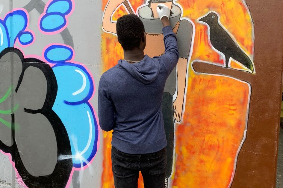 Ein junger Mann mit dunklen Haaren, schwarzer Hose und dunkelblauem Pullover sprüht mit einer weißen Spraydose an einer Graffiti-Wand. Er arbeitet an einer Figur mit hochgezogenem Schal und einem schwarzen Raben auf der Schulter, die auf einem leuchtend orangefarbenen Hintergrund gemalt ist. Am Boden liegen Holzkisten mit Spraydosen.