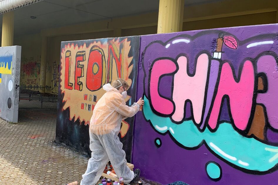 Eine Person in einem weißen Schutzanzug und Kapuze sprüht mit einer schwarzen Spraydose an einer Wand. Das Graffiti zeigt große, bunte Buchstaben mit einer lilafarbenen Hintergrundfläche. Daneben steht ein Schriftzug mit dem Namen „LEON“ in roten Buchstaben mit einem gelben Rahmen. Auf dem Boden liegen zahlreiche Spraydosen.