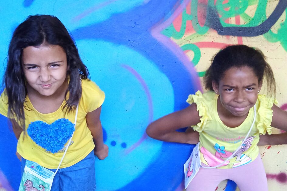 zwei Mädchen stehen vor einer bunten Graffiti-Wand. Beide tragen gelbe Oberteile und haben kleine Umhängetaschen. Sie machen spielerische, grimmige Gesichter und beugen sich leicht nach vorne.