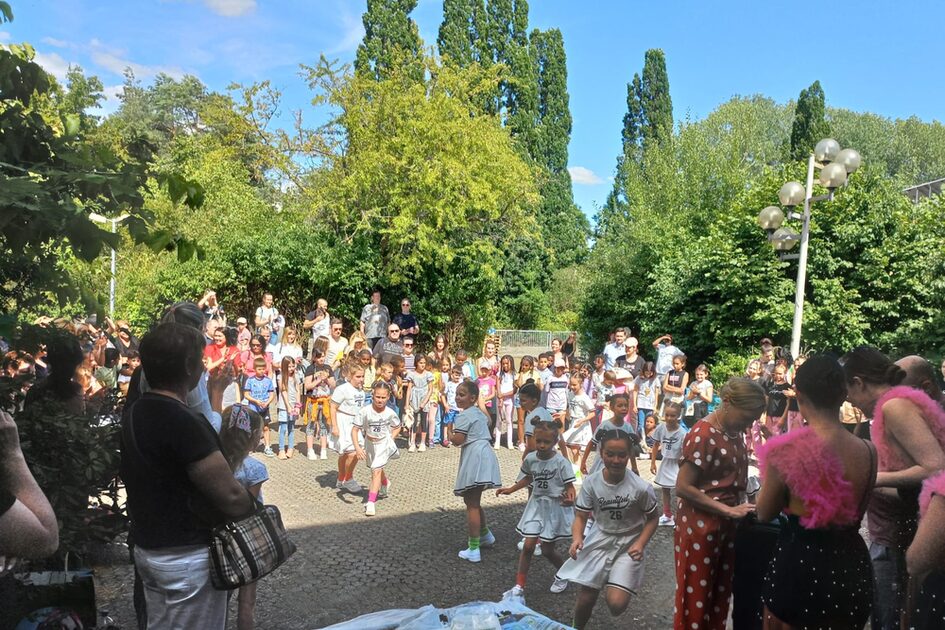 Eine große Gruppe von Menschen, darunter viele Kinder, versammelt sich im Freien an einem sonnigen Tag. Einige Kinder in weißen Sportoutfits laufen nach vorne, während Erwachsene am Rand zuschauen. Im Vordergrund sind Personen an einem Tisch mit Gegenständen.