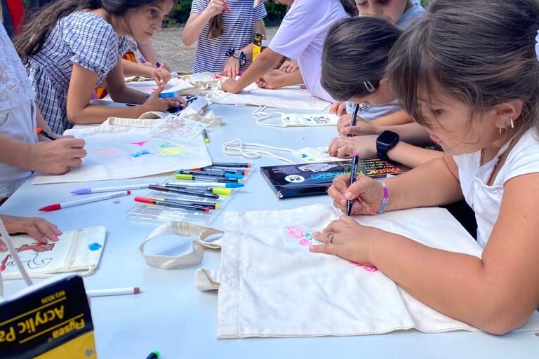 Kinder sind konzentriert beim Bemalen ihrer Stofftaschen. Der Tisch ist mit Farben, Stiften und Taschen gefüllt. Im Hintergrund stehen weitere Kinder und Erwachsene, einige mit Rucksäcken.