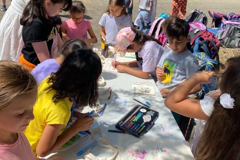 Mehrere Kinder sitzen an einem langen Tisch im Freien und bemalen Stofftaschen mit bunten Stiften. Verschiedene Malutensilien liegen verstreut auf dem Tisch.