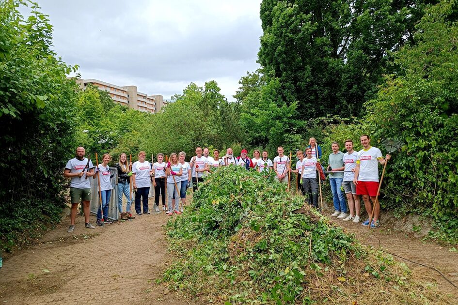 Eine Gruppe von etwa 20 Menschen steht auf einem gepflasterten Weg in einer grünen Umgebung. Sie tragen weiße T-Shirts mit rotem Schriftzug und halten Gartenwerkzeuge wie Rechen und Astscheren. Vor ihnen liegt ein großer Haufen geschnittener Zweige und Blätter. Im Hintergrund sind hohe Bäume und ein Wohngebäude zu sehen. Die Stimmung wirkt fröhlich und gemeinschaftlich.