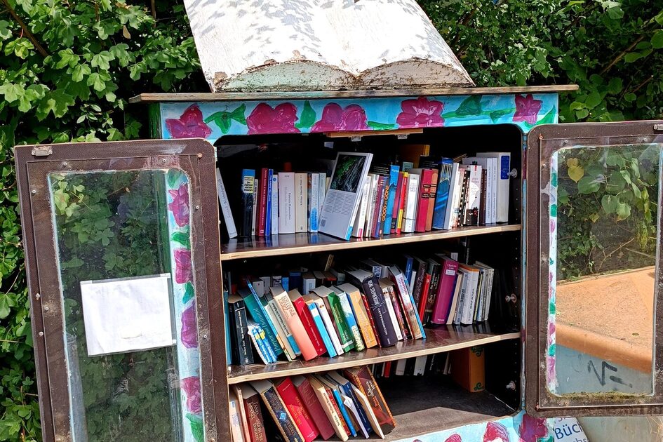 Offener Bücherschrank: Ein bunt bemalter Bücherschrank aus Holz mit Glasfenstern steht inmitten von grünen Sträuchern. Er ist gefüllt mit zahlreichen Büchern, die ordentlich auf Regalen stehen. Auf dem unteren Bereich des Schranks sind von Kindern gemalte Figuren mit roten Zipfelmützen zu sehen. Auf dem Dach befindet sich eine große, offene Buch-Skulptur, und darüber zeigt eine Wetterfahne die Himmelsrichtungen „N“ und „S“.