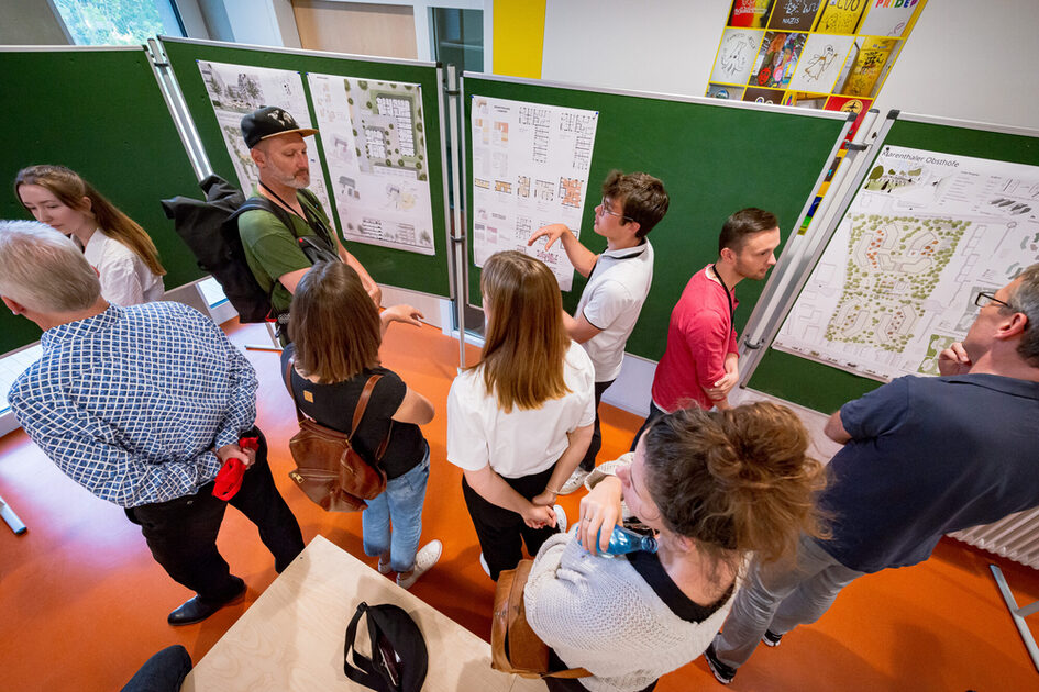 Mehrere Personen stehen in einem Raum mit orangefarbenem Boden und betrachten große grüne Pinnwände, an denen Architekturpläne und Entwürfe befestigt sind. Einige der Personen diskutieren aktiv über die Pläne, während andere zuhören oder sich Notizen machen. Die Gruppe besteht aus Männern und Frauen unterschiedlichen Alters, die lässige Kleidung tragen, darunter Rucksäcke und Umhängetaschen. Im Hintergrund sind bunte Kunstwerke oder Plakate an der Wand zu sehen. Eine Person im Vordergrund hält eine Wasserflasche.