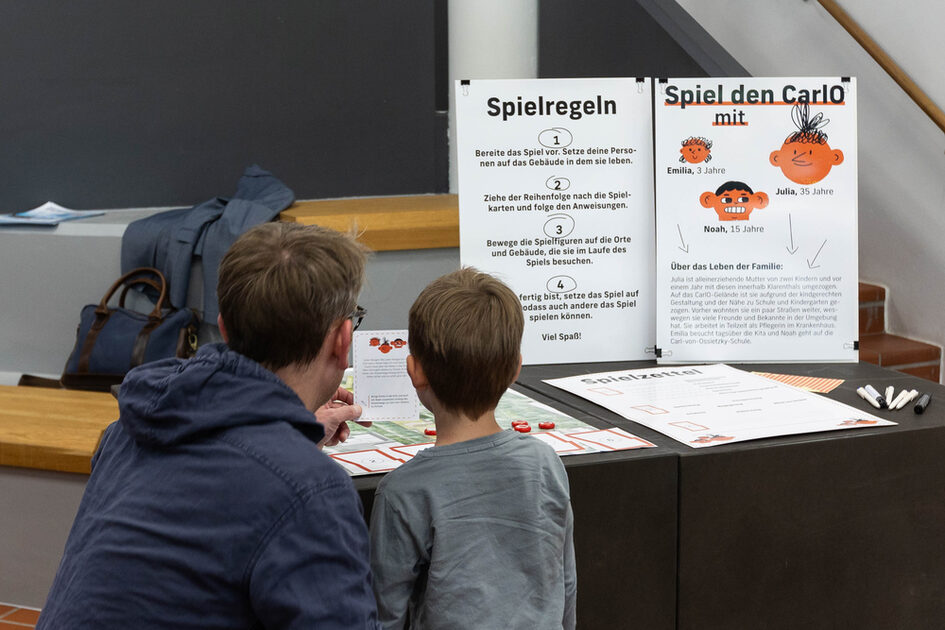 Ein Mann mit Brille und ein Kind mit kurzem Haar sitzen an einem Tisch und betrachten gemeinsam ein Lernspiel. Vor ihnen stehen zwei große Infotafeln mit Texten und Illustrationen. Eine Tafel trägt den Titel „Spielregeln“ und erklärt die Spielabläufe in nummerierten Schritten. Die andere Tafel stellt eine fiktive Familie mit den Namen Emilia, Julia und Noah vor. Auf dem Tisch liegen Spielkarten, Spielfiguren und ein Blatt mit der Überschrift „Spielzettel“. Im Hintergrund ist eine dunkle Tafel, eine Bank mit einer abgelegten Jacke sowie eine Tasche zu sehen.