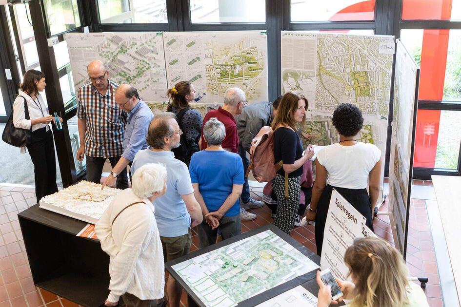 Eine Gruppe von Menschen steht in einem hellen Raum vor großen Plakaten mit Stadtplanungen und Architekturzeichnungen. Einige Personen studieren die Karten, während andere im Gespräch sind. Auf einem Tisch liegt ein dreidimensionales Modell eines Gebäudekomplexes. Im Vordergrund betrachtet eine Frau mit weißem Haar und beigem Pullover interessiert die Ausstellung. Eine Person macht ein Foto mit ihrem Smartphone. Die Fenster im Hintergrund lassen Tageslicht in den Raum.