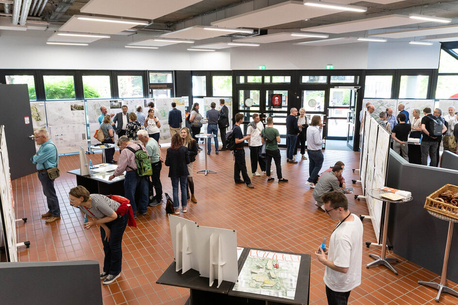 Eine Veranstaltung in einem modernen, offenen Gebäude mit hohen Fenstern und einer breiten Sitztreppe. Eine große Gruppe von Menschen sitzt auf den Stufen, während eine Person im Vordergrund an einem Rednerpult spricht. Auf den Tischen vor der Rednerin liegen verschiedene Broschüren, Zettel und Arbeitsmaterialien. Die Atmosphäre wirkt konzentriert und partizipativ.