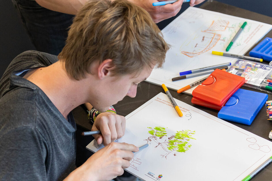 Eine junge Person mit kurzem Haar zeichnet konzentriert mit einem Filzstift auf einem Blatt Papier. Die Zeichnung zeigt einen Baum mit grünen Blättern. Um sie herum liegen viele bunte Stifte, zwei rote und ein blaues Stifteetui sowie weitere Zeichenblätter mit kreativen Skizzen. Eine zweite Person, deren Hand zu sehen ist, arbeitet ebenfalls an einer Zeichnung.