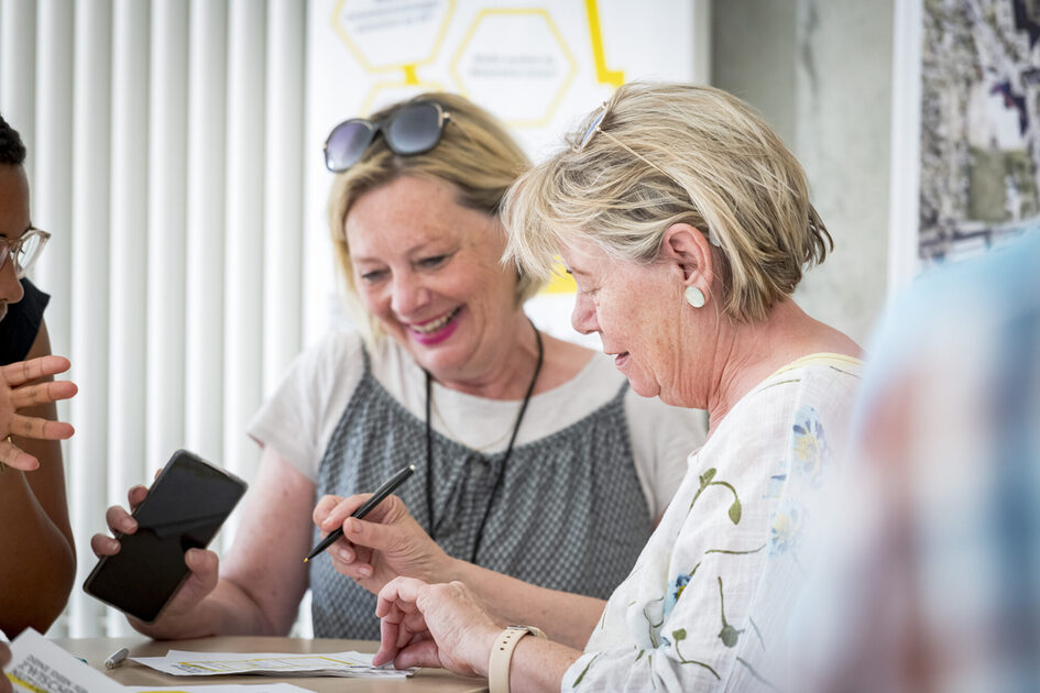 Zwei ältere Frauen stehen an einem Tisch und arbeiten gemeinsam an einem Dokument. Eine von ihnen hält einen Stift, während die andere lächelnd zuhört. Im Hintergrund sind Plakate mit gelben und weißen Elementen an der Wand angebracht. Die Atmosphäre wirkt freundlich und konzentriert.