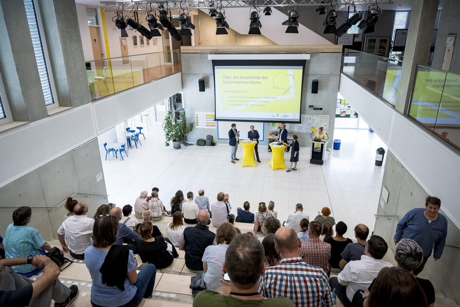 Eine große Veranstaltungshalle mit moderner Architektur und hohen Decken. Eine Gruppe von vier Personen steht auf einer Bühne mit zwei hohen, gelben Stehtischen. Eine Leinwand im Hintergrund zeigt eine Präsentation mit einem gelben Banner und dem Titel „Über die Geschichte der Experimentierräume“. Das Publikum sitzt auf breiten Stufen und hört aufmerksam zu.
