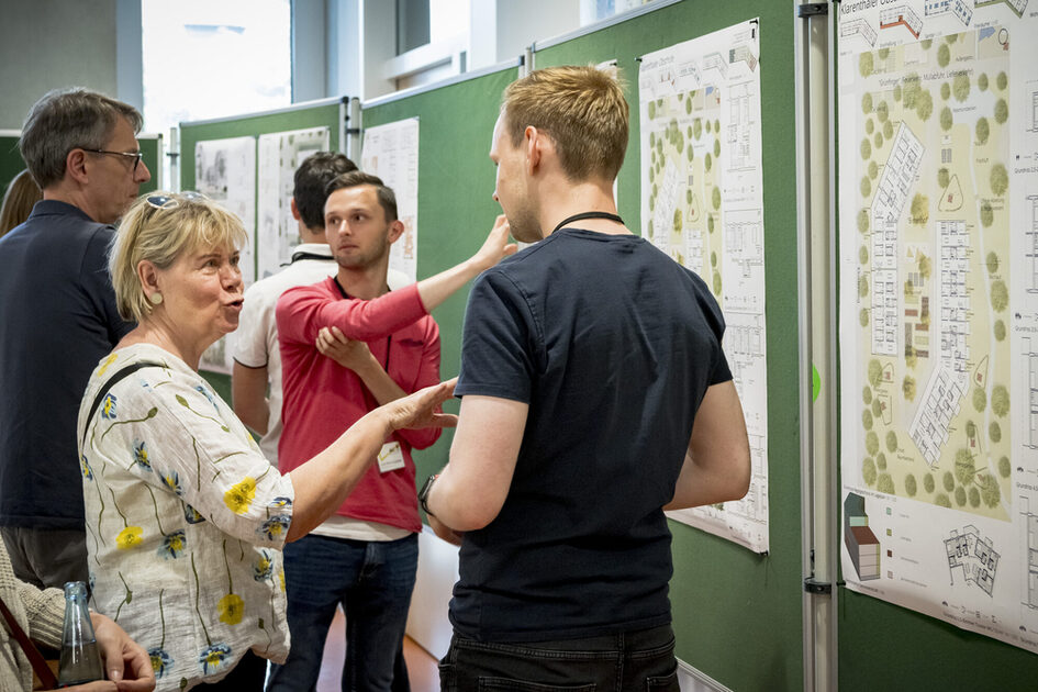 Eine Gruppe von Menschen steht vor großen, grünen Pinnwänden, auf denen Baupläne oder Karten befestigt sind. Eine ältere Frau mit kurzen, blonden Haaren in einer weißen Bluse mit gelben Blumenmustern spricht gestikulierend mit einem jüngeren Mann in einem dunklen T-Shirt. Eine weitere Person im Hintergrund zeigt auf einen bestimmten Punkt auf einem der Pläne.