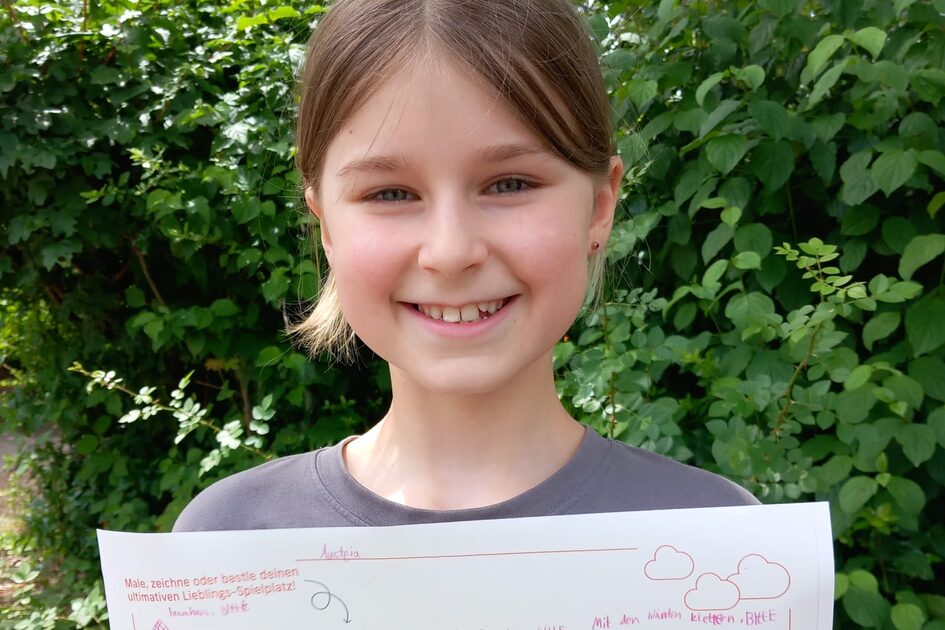 Ein junges Mädchen mit hellbraunen Haaren, die zu einem Mittelscheitel gekämmt sind, steht draußen vor einer grünen Hecke und lächelt in die Kamera. Sie trägt ein graues T-Shirt. In ihren Händen hält sie ein Blatt Papier mit einer selbstgezeichneten Skizze ihres Traum-Spielplatzes. Die Zeichnung ist in rot und pink gehalten und enthält verschiedene Spielgeräte, darunter eine Rutsche, ein Klettergerüst und ein Karussell. Das Blatt trägt den Titel „Male, zeichne oder bastle deinen ultimativen Lieblings-Spielplatz!“ und enthält Logos von GWW und der Stadt Wiesbaden.