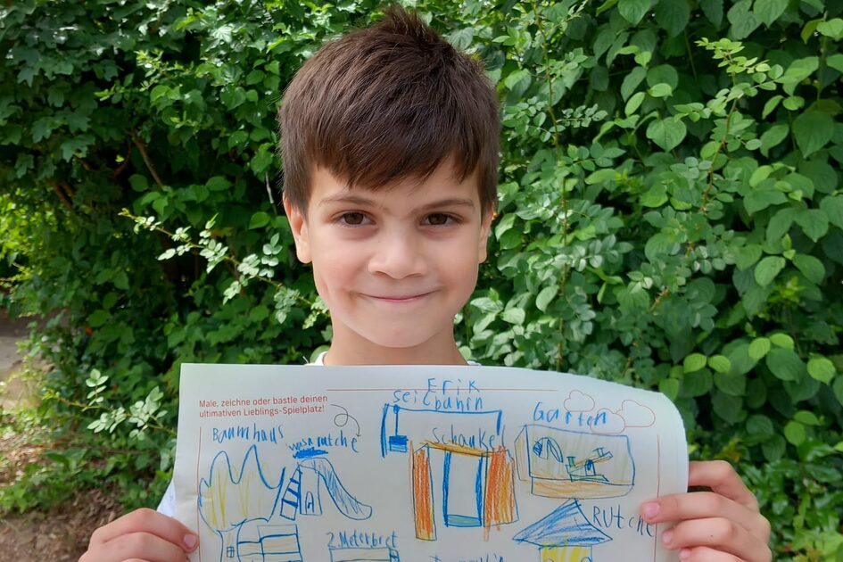 Ein Junge mit kurzen braunen Haaren steht vor einer dichten grünen Hecke und schaut freundlich in die Kamera. Er trägt ein weißes T-Shirt mit bunten Mustern. In seinen Händen hält er eine Zeichnung seines idealen Spielplatzes, die mit blauen, orangen und grünen Buntstiften gestaltet wurde. Zu sehen sind ein Baumhaus mit Rutsche, eine Schaukel, eine Wippe und ein Trampolin. Der Name „Erik“ ist oben auf dem Blatt notiert. Auch dieses Blatt enthält die Logos von GWW und der Stadt Wiesbaden.