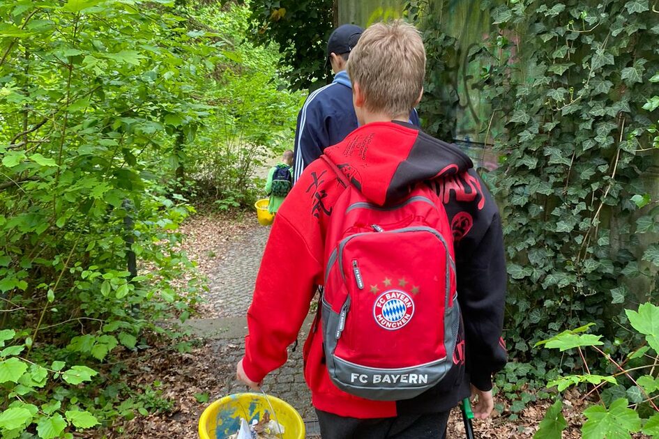 Ein Junge mit einem roten FC-Bayern-Rucksack läuft auf einem schmalen, mit Laub bedeckten Weg durch eine grüne, bewachsene Umgebung. Er hält einen gelben Eimer mit gesammeltem Müll in der rechten Hand und eine Müllzange in der linken Hand. Vor ihm geht eine weitere Person mit einer dunklen Jacke und einer schwarzen Mütze. Eine hohe Betonwand, teilweise mit Graffiti bedeckt, ist rechts im Bild zu sehen.