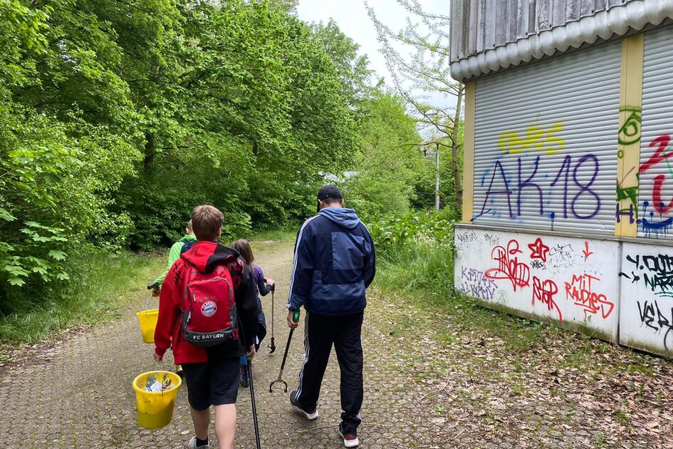 Mehrere Personen laufen auf einem gepflasterten Weg an einem mit Graffiti besprühten Gebäude vorbei. Der Junge mit dem roten FC-Bayern-Rucksack ist erneut zu sehen, ebenso eine Person in einer blauen Jacke. Alle tragen Müllzangen und Eimer mit Müll. Der Weg ist von hohen, grünen Bäumen gesäumt.