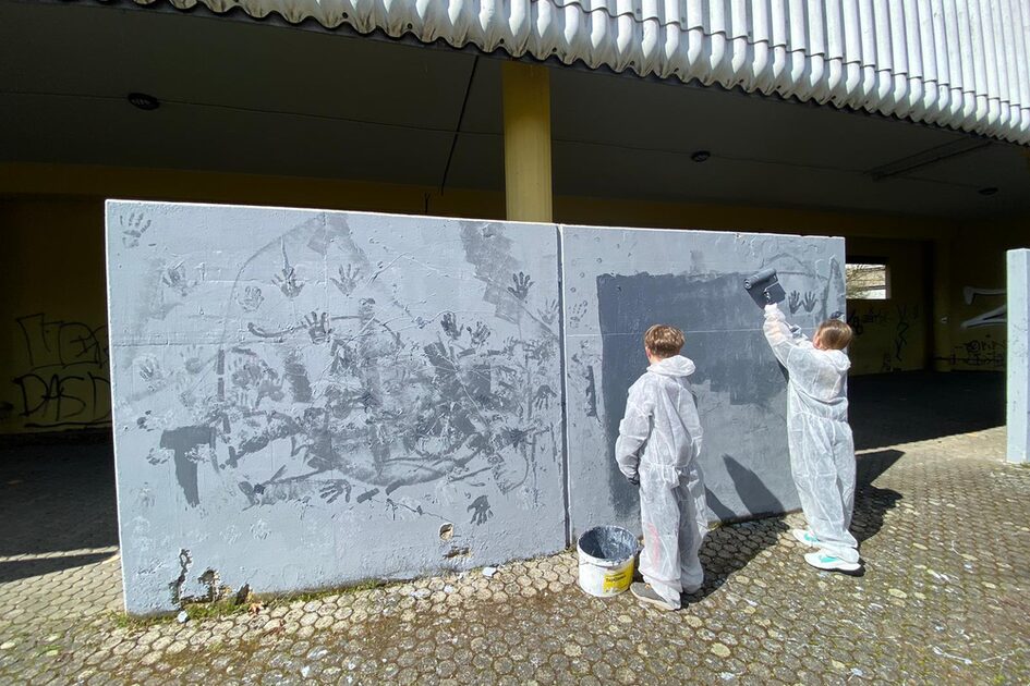 Zwei Kinder in weißen Schutzanzügen bemalen eine Betonwand mit grauer Farbe. Die Wand ist bereits mit vielen schwarzen Handabdrücken bedeckt. Ein Kind trägt die Farbe mit einer großen Rolle auf, während das andere zusieht. Vor ihnen steht ein schwarzer Farbeimer.