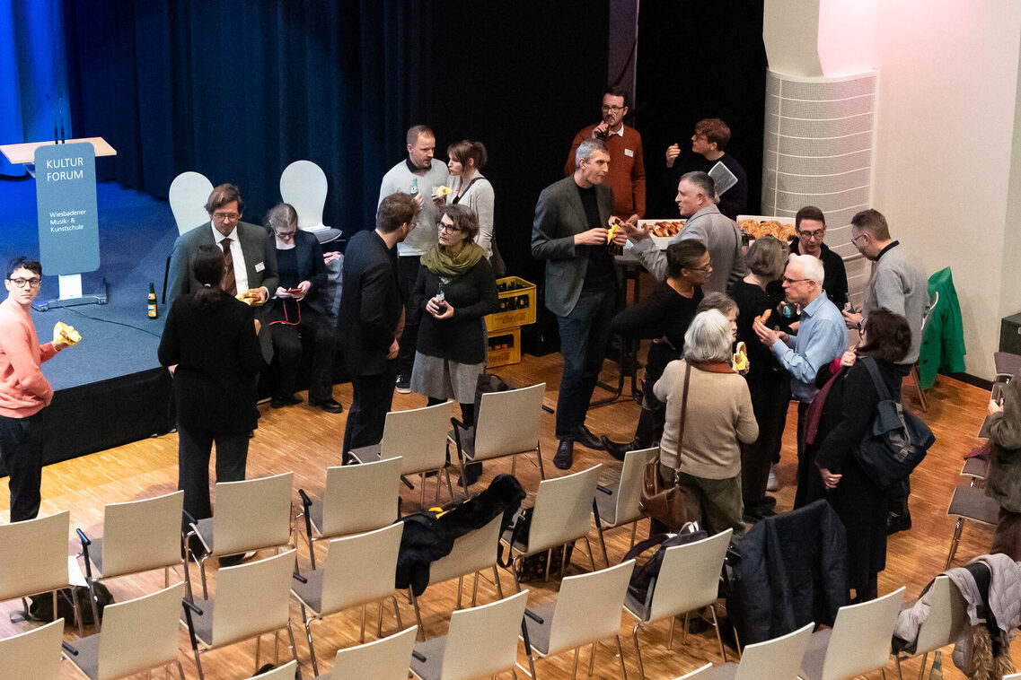 Die Teilnehmenden stehen in einem bestuhlten Saal vor der Bühne und unterhalten sich.