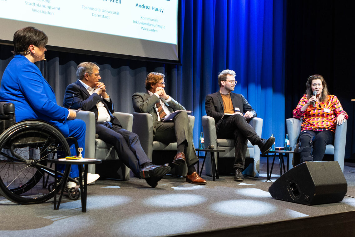 Drei Männer und eine Frau sitzen in Sesseln auf einer Bühne, während die Frau etwas in ein Handmikrofon spricht. Eine weitere Frau sitzt auf der Bühne im Rollstuhl.