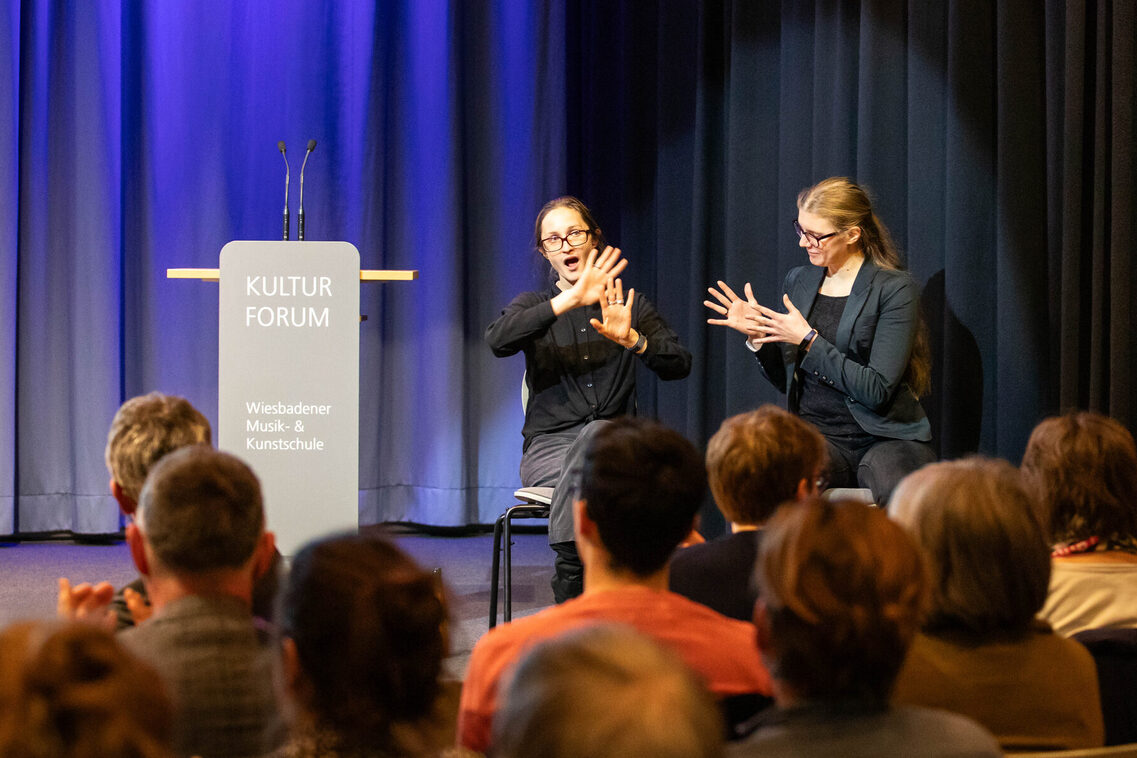 Zwei Frauen sitzen auf der Bühne und Übersetzen das gesprochene Wort in Gebärden. Im Vordergrund sieht man die Köpfe der Teilnehmenden.