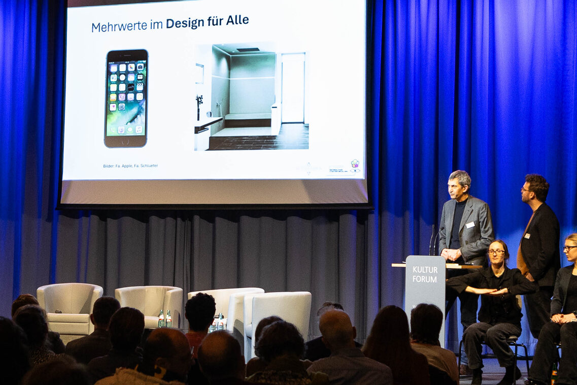 Zwei Männer stehen auf der Bühne an einem Pult. Vor ihnen sitzen zwei Frauen. Im Hintergrund zeigt eine Präsentation das Bild eines Handys und eines Badezimmers.