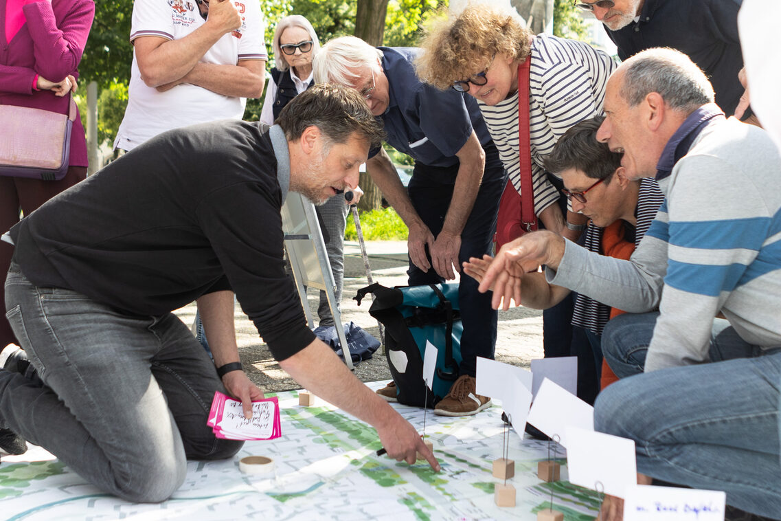 Eine Gruppe von Frauen und Männern diskutiert. Sie knien und beugen sich über ein Plakat auf dem ein Mann der Gruppe etwas zeigt.