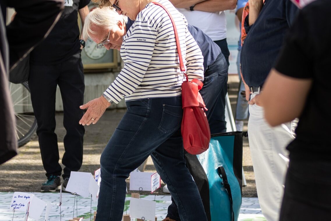 Eine Gruppe von Frauen und Männern steht auf einem großen Plakat. Eine Frau zeigt einem Mann darauf etwas mit ihrem Fuß und ihrer Hand.